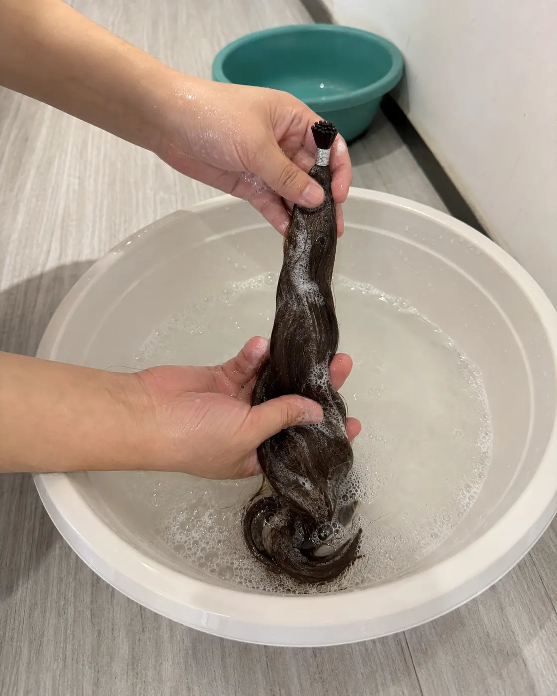 Hands washing a dark-brown Remy human hair extension sample with shampoo foam in a clear water basin during an Alpha Hair wash test
